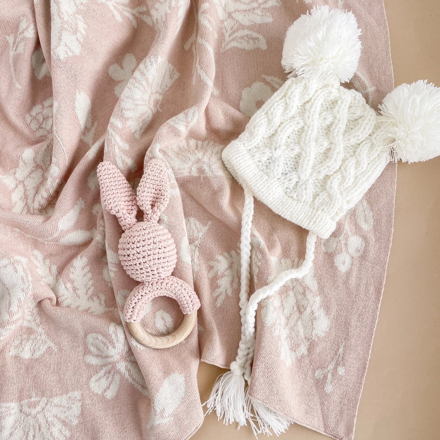 Pink knitted teething ring and white knit hat with pom-poms on a pink floral blanket.